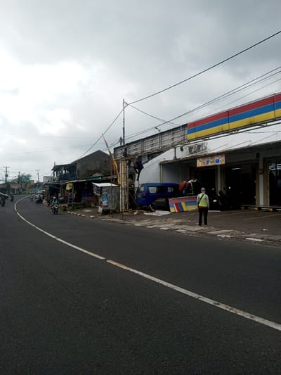 A street scene on an overcast day. Motorbikes travel along an asphalt road marked with a white line. A building with a red, blue, and yellow sign stands on the right. Two workers are on a ladder near a blue van, appearing to install or repair the sign. A person in a yellow vest stands nearby, observing. Other buildings and shops line the street beyond. Overhead, power lines crisscross the sky.