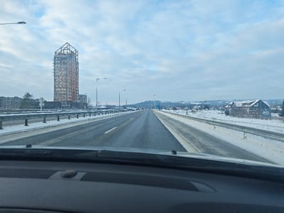 The image depicts a winter highway scene from the interior perspective of a vehicle, looking forward. The foreground shows the dark gray dashboard and windshield base. Ahead, a multi-lane highway, likely E6, extends into the distance, with clear, dark asphalt lanes bordered by snow-covered shoulders and median strips. Guardrails run along both sides of the highway. Numerous streetlights are positioned along the road.

To the left of the highway, the prominent Mjøstårnet, a tall, multi-story wooden skyscraper, stands against a cloudy, light blue sky. Its distinct wooden frame is visible at the top, and numerous windows line its facade. Adjacent to Mjøstårnet, other lower-rise buildings and snow-covered ground are visible.

To the right of the highway, beyond the guardrail and snow-covered terrain, a building with a gabled roof, possibly a hotel or restaurant (a sign partially displays "SCANDIC"), is present. Further in the distance on the right, a tall pole bearing a prominent yellow "M" logo, indicative of McDonald's, is visible. The entire landscape, including distant hills and structures, is covered in a layer of snow, indicating a winter setting. The sky is overcast with scattered clouds. The scene is situated in Brumunddal, Norway.