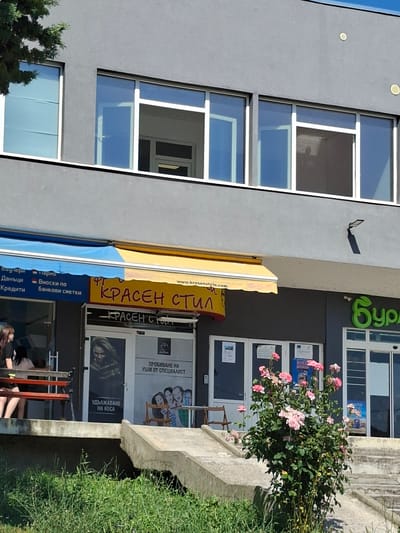 The image depicts a commercial building with a gray facade and multiple windows. The building is situated in Aksakovo, Bulgaria. In the foreground, there is a ground-level entrance area with awnings over storefronts. One awning is yellow and branded with "КРАСЕН СТИЛ" (Krasen Stil). Below this awning, a storefront displays text in Bulgarian, including "ПРОБИВАНЕ НА УШИ ОТ СПЕЦИАЛИСТ" (Ear Piercing by Specialist) and "УДЪЛЖАВАНЕ НА КОСА" (Hair Extension). A poster of a woman with long hair is visible on a glass door. To the right of this storefront, another business, likely a pharmacy or health-related store, is indicated by a large green leaf logo and the partial word "Бур" (Bur), suggesting "Бурбон" (Bourbon) or a similar brand. A bush with pink roses is planted in front of the building, and a concrete ramp leads to the entrance level. Two young women are seated on a bench to the left of the entrance. The environment appears to be a sunny day.