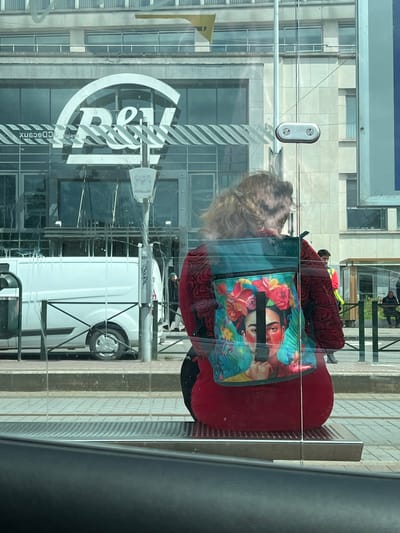 The image shows a woman wearing a red jacket and a backpack featuring a Frida Kahlo design, standing behind a glass barrier. The background reveals a street scene in Saint-Josse-ten-Noode - Sint-Joost-ten-Node, Belgium. A white van is parked on the left, and a building with the "R&V" logo is visible behind the woman. The glass barrier is reflective, showing the woman and the surroundings, with some smudges and streaks on its surface. The ground is paved with tiles and a metal grate. There are other people visible in the background, including a man in a yellow vest. The overall setting appears to be a public transportation stop or a busy urban area.