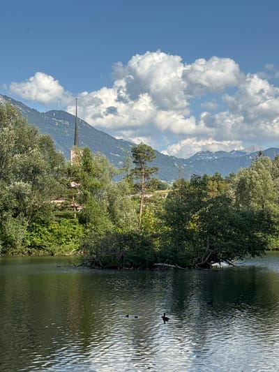 The image depicts a serene lake with a small island covered in dense green foliage. Several ducks are visible swimming on the water's surface. In the background, a tall, slender church spire with a pinkish hue rises above a lush canopy of trees. Beyond the trees, majestic mountains with rugged peaks, some dusted with snow, form a dramatic backdrop under a bright blue sky dotted with fluffy white clouds. The overall scene suggests a tranquil natural setting with architectural elements integrated into the landscape. While the exact location within Buchs (SG), Switzerland, isn't explicitly identifiable from the image alone, it captures the characteristic beauty of the Swiss Alps and its surrounding environments.