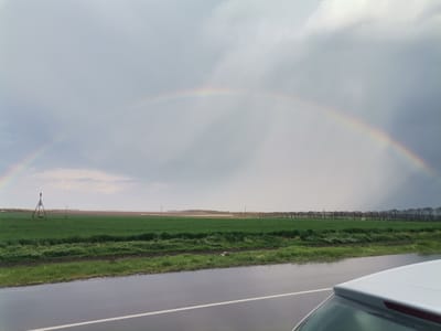A wide landscape photograph captures a distinct rainbow arching across a cloudy sky over a rural scene. In the foreground, a wet asphalt road, marked with a white lane line, occupies the lower third of the image. The upper part of a white car is visible on the far right, suggesting the photo was taken from inside or beside a vehicle. A narrow ditch separates the road from a large, verdant green field that fills the midground. Within the field, an A-frame irrigation sprinkler system tripod is visible on the left, and several utility poles are scattered. A dense line of trees marks the far edge of the field, with lighter fields or barren land beyond them. The sky is predominantly overcast with varying shades of gray clouds, but the rainbow is clearly visible. The wet road surface indicates recent rainfall. The scene depicts a daytime view in Slaveevo, Bulgaria. No visible text is present.