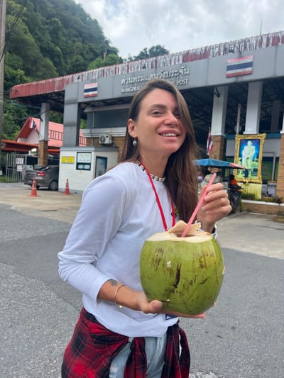 A woman with long brown hair, wearing a white long-sleeved shirt, ripped blue jeans, and a red and black plaid shirt tied around her waist, stands outdoors holding a large, green coconut with a pink straw. She is smiling and looking at the camera. Behind her is a building with Thai writing on it, identified as "WANGPRACHAN POST." The building features a Thai flag and a framed portrait of a man in uniform. The surrounding environment appears to be a street with trees and hills in the background. The scene is set in Tambon Wang Prachan, Thailand.