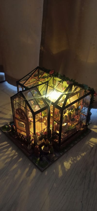 The image shows a miniature greenhouse diorama, likely a craft or hobby item. The greenhouse is made of glass panels and dark framing, adorned with small green vines and colorful faux flowers. Inside, it is illuminated by a bright light source, revealing various miniature objects such as plants, vases with flowers, and possibly small figurines. The diorama is placed on a marble-like surface, with light casting dappled shadows across it, suggesting the lighting is coming from an external source. The scene appears to be indoors, possibly in a home environment, given the flooring and ambient lighting. There is no visible text in the image. The overall impression is one of intricate detail and a charming, whimsical miniature world.