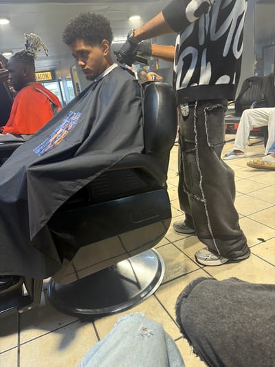 This image captures a barbershop scene in Blouberg, South Africa. In the foreground, a client with dark, curly hair sits in a black barber chair, covered by a black barber cape. He is looking downwards as a barber attends to his hair. The barber, standing behind the client, wears a black and white patterned long-sleeved top, distressed black jeans with visible fraying and white stitching, and black gloves. He is actively using hair clippers on the client's head.
The tiled floor, a mix of light beige and brown squares, is visible beneath the chairs. A round, metallic tray for hair clippings is positioned on the floor near the client's feet. In the mid-ground, another client is visible, seated in a separate barber chair, covered by an orange cape, with distinctive light-colored dreadlocks. Further in the background, another person is partially visible, wearing light grey pants and patterned slippers. The interior of the salon is well-lit, with light fixtures visible. A yellow sign in the background clearly displays the word "SALON." The black barber cape in the foreground features a small, blue and orange emblem on its lower front. The overall scene depicts a standard haircut in progress within a busy salon environment.
