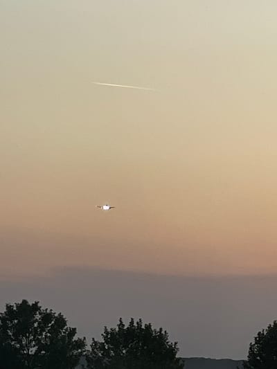 The image, when viewed in correct landscape orientation, depicts a hazy sky above Otopeni, Romania, likely during dawn or dusk. The sky presents a gradient of colors, transitioning from a lighter, pale beige or soft orange hue on the left to a darker, greyish tone on the right. Along the right edge of the frame, dark silhouettes of tree foliage are visible, suggesting a natural or suburban setting.

Two distinct elements are observed in the sky:
1.  On the upper left, a bright, thin, white streak extends diagonally across the hazy sky, indicating a luminous object in motion, possibly a contrail from an aircraft, a meteor, or another fast-moving light source.
2.  Near the center, a smaller, indistinct object is visible, characterized primarily by a prominent bright light emanating from its bottom. The dark shape above the light is unclear due to distance and blur, but it could suggest a distant aircraft, helicopter, or drone.

The overall atmosphere is diffuse and muted, indicative of atmospheric haze or partial cloud cover. There is no human activity or visible text in the image.