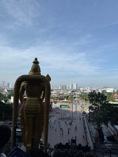 This image, taken from an elevated perspective, showcases a magnificent golden statue overlooking an urban panorama under a clear sky. The prominent subject is a large, intricately detailed golden statue of a deity, possibly Lord Murugan, positioned on the right side of the frame. The statue wears a tall, ornate headdress and features a textured, snake-like garland or embellishment draped around its shoulders and arms.

Below the statue, a wide, paved plaza stretches out, populated by numerous people walking and gathering, suggesting a public or religious site. Beyond the plaza, a bustling city unfolds with an array of buildings, roads, and various vehicles, including a green bus and parked cars. Trees and foliage are interspersed throughout the urban landscape, and utility poles with power lines are visible in the background.

The sky is a bright blue with scattered, wispy clouds, indicating fair weather. The time of day appears to be daytime, likely morning or afternoon, given the consistent natural lighting. On a distant billboard within the cityscape, the word "POWERCARE" is clearly visible. The overall scene suggests a vibrant cultural or religious landmark, such as the Batu Caves in Malaysia, within an active urban environment.