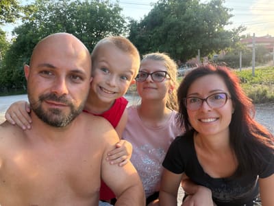 A group of four people, two adults and two children, are taking a selfie outdoors. The adult male in the foreground is shirtless and bald, with a beard. A young boy with blond hair and a wide smile is to his right, wearing a red shirt. Next to him, a young girl with glasses and light brown hair is smiling, wearing a pink shirt with a floral print. An adult female with dark hair and glasses is on the far right, also smiling and wearing a black t-shirt. They appear to be in a semi-rural area, with trees and a fence visible in the background. The lighting suggests it is late afternoon or early evening. The location is identified as Dolni Dabnik, Bulgaria.