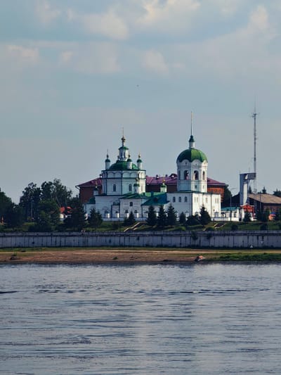The image captures a panoramic view of the Yeniseysk Theological Seminary complex in Yeniseysk, Russia, situated on the right bank of the Angara River. The main architectural feature is a white church with multiple green domes, characteristic of Russian Orthodox architecture. To the right of the church, a tall communication tower is visible. The complex is bordered by a concrete embankment along the river. The water in the foreground is rippled, suggesting a gentle current. The sky above is partly cloudy with a soft blue hue. The scene is bathed in natural light, indicating it was taken during the daytime, likely under a clear or partially overcast sky.