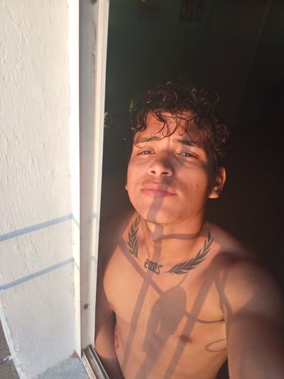 This is a medium shot, taken from a slightly low angle, of a young man with curly dark hair, looking directly at the camera. He is shirtless, exposing his chest and shoulders, and has a tattoo on his neck that appears to be a laurel wreath with the number "2005" next to it. The man's skin is illuminated by warm, golden light, casting shadows across his face and body, suggesting it is either sunrise or sunset. He is standing in what appears to be a doorway or opening, with a textured white wall on the left side of the frame that has a shadow pattern cast upon it, indicative of sunlight filtering through blinds or a window frame. The background behind the man is dark and out of focus, with a hint of greenery visible. The image is likely taken in La Guardia, Venezuela, given the mention of its location.