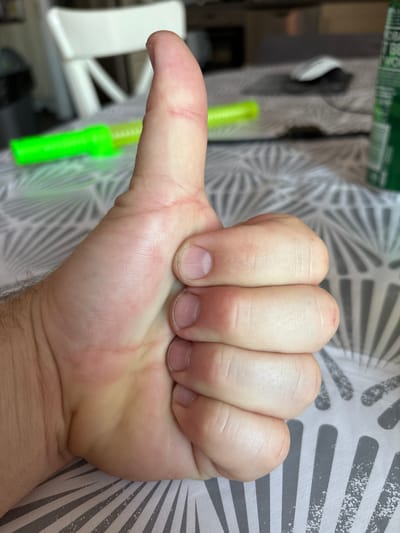 This is a close-up, eye-level photograph of a person's right hand giving a thumbs-up gesture. The hand is positioned in the foreground, obscuring most of the background. The thumb is extended upwards, and the fingers are curled inwards, forming a fist. The skin on the back of the hand and fingers appears lightly tanned and has visible creases and lines. The fingernails are short and appear clean. The background is out of focus, showing a patterned tablecloth, a white chair, and some objects that are not clearly identifiable, including a green item that may be a can or bottle and a green cylindrical object. The overall lighting suggests an indoor setting, possibly a dining area or a kitchen. The image does not contain any elements that would allow for the determination of its location in the city of Cork, Ireland.