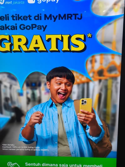 The image shows a digital advertisement for MRT Jakarta and GoPay. The advertisement features a young man smiling and holding a yellow smartphone. The background shows a blurry image of a train, likely on the MRT Jakarta line. Text on the advertisement reads "Beli tiket di MyMRTJA pakai GoPay GRATIS". The advertisement is likely displayed in a public space in South Jakarta, Indonesia, such as an MRT station or a shopping mall. The young man appears to be excited about the promotion, which offers free tickets when purchased using GoPay. The overall tone of the advertisement is positive and engaging.