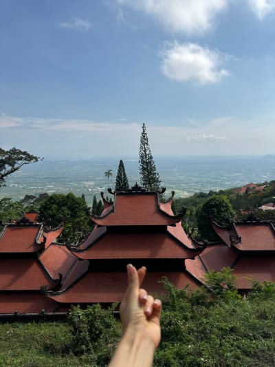 A person's hand with neatly manicured nails is positioned in the foreground, making a finger heart gesture. Behind the hand, a multi-tiered traditional Vietnamese temple with red tiled roofs and dark, ornate eaves dominates the mid-ground. Lush green foliage, including trees and bushes, surrounds the temple structures. In the background, a sprawling landscape unfolds, featuring a vast, green valley dotted with buildings and agricultural fields, stretching towards a hazy horizon under a bright blue sky with scattered white clouds. The scene is likely filmed in Thuận Nam, Vietnam, given the architectural style and the typical landscape of the region.