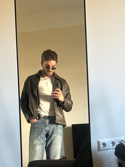 An indoor scene in Bertrange, Luxembourg, captures a male individual taking a mirror selfie. The subject is reflected centrally in a full-length mirror with a black frame. He wears a dark brown leather jacket over a light-colored, three-button Henley shirt, and light blue denim jeans. His right hand holds a dark-colored smartphone, oriented to capture his reflection. His left hand is positioned within his left jeans pocket. The subject has medium-length brown hair, light facial stubble, and wears small, oval, tinted sunglasses. The wall reflected behind the individual is a plain, light beige. To the immediate right of the mirror, a white, textured wallpapered wall is visible, featuring a white triple Schuko-style power outlet with two plugs inserted. A dark, indeterminate object, possibly furniture, is partially visible in the lower right foreground.