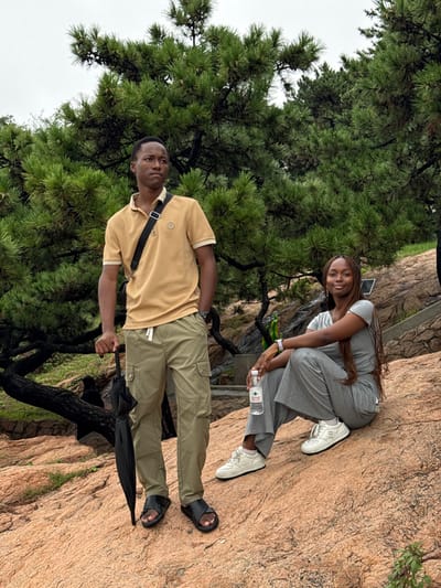 This image depicts an outdoor scene in Shinan District, China, featuring two individuals amidst a rocky landscape with pine trees under an overcast sky. On the left, a male subject stands, positioned in a three-quarter profile facing right. He wears a tan polo shirt with a lighter collar, khaki cargo pants with a white drawstring, and black cross-strap sandals. A black cross-body bag is worn across his chest. His left hand holds a closed black umbrella, while his right is in his pocket. On the right, a female subject sits on a large reddish-brown rock, facing slightly left with her gaze directed towards the viewer. She is dressed in a grey short-sleeved top and matching wide-leg pants, paired with white sneakers. A silver-colored watch is visible on her left wrist. She holds a clear plastic water bottle with a white label, bearing Chinese characters, in her right hand. The immediate ground consists of varied reddish-brown rock formations, sloping gently upward. In the mid-ground, behind the female subject, a small, dark green decorative element is partially visible. The background is densely populated with mature pine trees, displaying dark green needles and visible dark, gnarled branches.