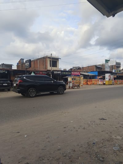 The image depicts a roadside scene in Gampongpaya, Indonesia. In the foreground, a dark-colored SUV is parked on the right side of a paved road. The road surface is dusty and shows signs of wear, with scattered small stones and debris. To the left of the SUV, another dark-colored vehicle is partially visible.
Behind the vehicles, a row of businesses and street food stalls lines the road. These structures appear to be a mix of permanent buildings with brick walls and corrugated metal roofs, and temporary stalls. Signs are visible above the businesses, including one for "AMRI STYLE" and another advertising "WM BOUTIQUE". Several food carts are present, one with a prominent sign for "WIN DIMSUM" and another displaying "HARFI PIZZZA". Orange plastic chairs are stacked near one of the stalls.
The environment appears to be somewhat overcast, with a cloudy sky dominating the upper portion of the image. Utility wires are strung across the sky. The overall impression is of a bustling commercial area along a main road in a town or city.