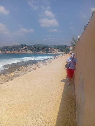 A person stands next to a tan wall, looking at their phone. They are wearing red shorts, a light blue shirt, a beige cap, and a dark bag slung across their body. The person is on a gravel path that runs alongside a rocky shoreline. Waves crash against the rocks, and the Mediterranean Sea is visible in the background. A town with terracotta roofs is nestled on a hill overlooking the water. This scene is located in Bandol, France, on the coast.