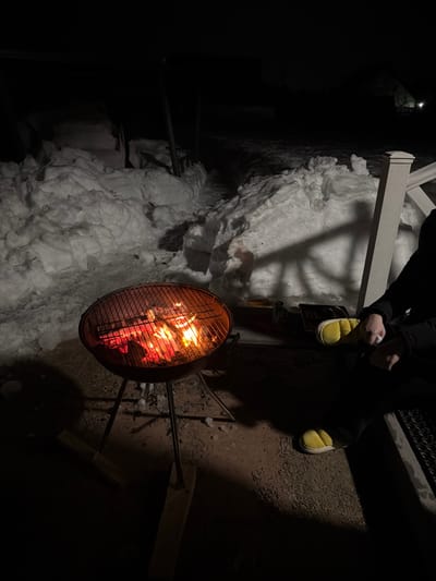 This nighttime image, captured in Ветеран Снт, Russia, depicts an outdoor scene dominated by a lit barbecue grill and a partially visible individual. In the foreground, a black kettle-style charcoal grill stands on three legs, filled with glowing red and orange embers and flames, radiating heat. A metal grate covers the top of the grill. To the right, a person is seated, visible from the torso down. They wear dark pants and bright yellow, soft slippers. Their right hand is visible, holding a small white object, possibly a napkin or tissue. Directly behind the person is a white railing post. A faint shadow of a lattice or railing structure is cast onto the snow-covered ground behind the person. The ground immediately surrounding the grill and the seated person is dark, bare earth or gravel. Beyond this area, the environment is heavily covered in deep snowdrifts, with a narrow, cleared path visible winding into the dark background. The scene is illuminated primarily by the intense light from the grill fire, contrasting with the overall darkness.