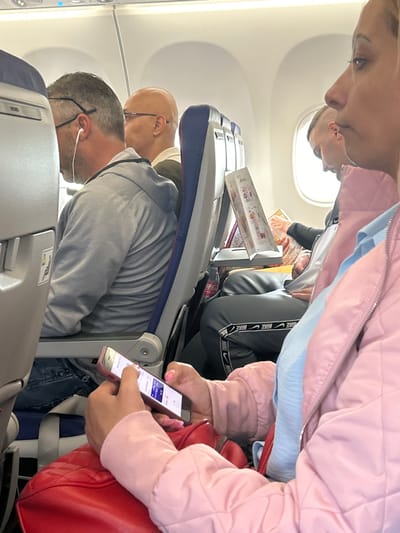 The image displays the interior of an airplane cabin, appearing rotated clockwise by 90 degrees, suggesting a perspective from a passenger's seat. Bright light from the windows indicates daytime.

In the foreground, a person is wearing a pink quilted jacket over a light blue garment. Their hand, with pink manicured nails, holds a light-colored smartphone displaying a screen with white, purple, and blue interface elements. A red shoulder bag or purse rests beside them.

Further into the cabin, several other passengers are visible. A man with short hair, wearing glasses and a gray hoodie, is seated in front. He has white wired earphones in his ears. Behind him, another bald man with glasses and an earring is partially visible. Further back, a passenger is wearing black sweatpants adorned with "NIKE" branding along the side stripe. Below the seats, a partially visible Coca-Cola bottle can be seen.

One of the tray tables is lowered, holding an in-flight magazine or menu, where the text "Hot Drinks" is discernible on its cover. A clear reflection of a person's head is visible in the airplane window. The scene depicts a calm flight, possibly originating from or arriving at Dortmund, Germany.