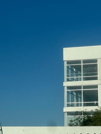 The image displays a modern, multi-story building under a clear, unobstructed blue sky in La Venta, Mexico. The building occupies the right portion of the frame, presenting a light gray or off-white facade. It features at least three visible levels, each characterized by large, rectangular windows with white frames that reflect the surrounding blue sky. The lowest visible section of the building and its corresponding windows are partially obscured by green foliage from trees or bushes at the bottom right. A light-colored, horizontal wall or parapet extends across the bottom edge of the frame. No human figures, animals, or overt activities are present within the scene.