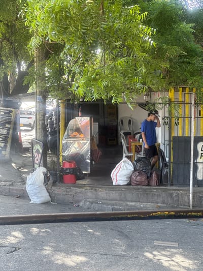 In Perímetro Urbano Barranquilla, Colombia, a young male wearing a blue t-shirt and black shorts stands on a sidewalk near a food stall. He is wearing a baseball cap and appears to be touching his forehead. In front of him are several large trash bags, two brown and one white with red stripes, and a black plastic bag. To his left is a glass-fronted food display case filled with what appears to be fried chicken. Behind the food stall, white chairs and tables are visible, along with yellow and white striped poles. The scene is set under a canopy of lush green trees, with sunlight dappling through the leaves. The street in the foreground is asphalt and shows some wear and tear. A metal pipe runs horizontally across the lower part of the frame.