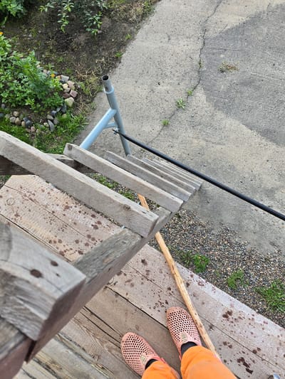 The image depicts a close-up, elevated view of a wooden ladder and a person's legs wearing orange trousers and pink perforated clogs, standing on wooden planks. The ladder appears weathered and is positioned outdoors, with a gray concrete path running alongside it. A portion of a garden with green plants and rocks is visible to the left. A metal pipe, possibly part of a structure or support, extends from the upper left of the frame diagonally across the concrete. The scene appears to be in a backyard or outdoor area. No specific landmarks identifiable as being in Ермолаево, Russia are visible.