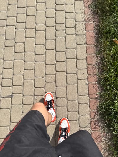 The image shows a person's legs and feet walking on a cobblestone path next to a grassy area. The person is wearing black shorts with red detailing and white and red sneakers with black laces. The cobblestone path is made of rectangular and square pavers. To the right of the path is a strip of red pavers, followed by a patch of green grass. The scene appears to be outdoors on a sunny day, as indicated by the shadows cast on the path and grass. The specific location is on a path in the city of Białystok, Poland, likely a park or a pedestrian walkway.