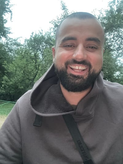 The image is a close-up, selfie-style shot of a man with a shaved head and a beard. He is smiling broadly, showing his teeth. He is wearing a dark grey hooded sweatshirt with the hood up. A black strap with some visible lettering is visible across his chest. The background consists of out-of-focus green trees and a bright, overcast sky. Based on the provided information, the location is the city of Lytkarino, Russia.