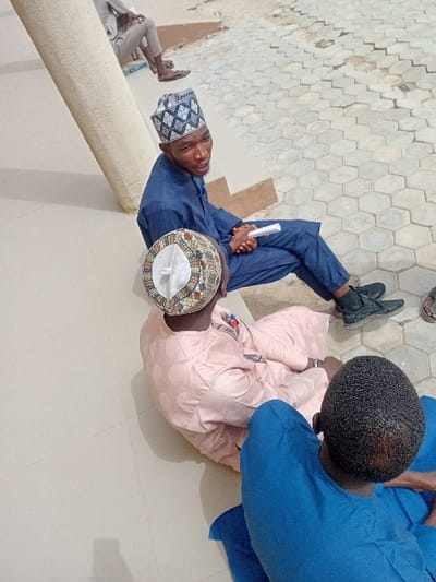 The image shows an outdoor gathering of four men in the city of Kwazailewa, Nigeria. In the foreground, a man with short, dark hair, wearing a blue traditional tunic, sits on a light-colored tiled surface with his back to the camera. To his right, another man, dressed in a light pink traditional garment and a multi-colored cap, sits facing left. He has a watch on his left wrist. Ahead of them, a third man, wearing a dark blue traditional outfit and a black and white patterned cap, sits on a light-colored step or raised platform. He holds a white rolled document in his hands and is positioned as if speaking, with his mouth slightly open. He wears black sneakers. In the upper background, a fourth individual is partially visible, seated on a higher step, wearing light-colored trousers and sandals. The ground features smooth, light-colored tiles in the foreground and hexagonal pavers in the mid-ground. A light-colored cylindrical pillar is visible on the left side of the frame.