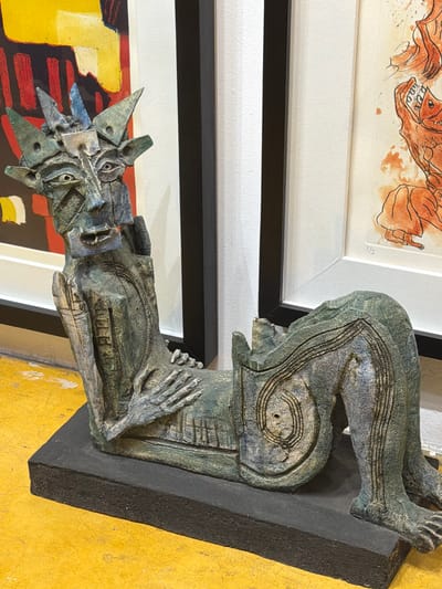 A contemporary sculpture rests against a yellow wall, in Oaxaca City, Mexico. The sculpture is primarily grey-green, mounted on a black base, and depicts a distorted figure with multiple eyes and a crown-like structure on its head. The body is stylized with carved lines. The sculpture is positioned in front of two framed art pieces hung on a white wall. The artwork above is a drawing in shades of orange and brown, while the piece below has sections of yellow, brown, and red. All these objects are in an indoor setting.