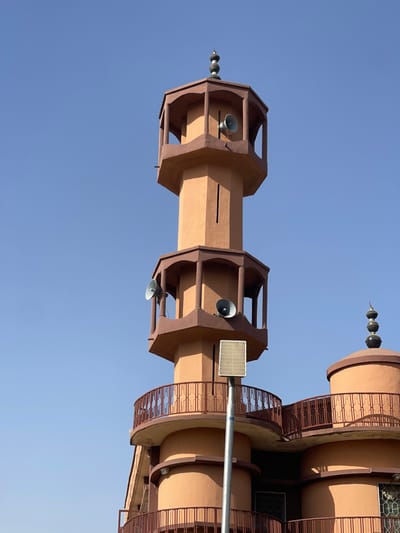 This image captures a portion of a mosque structure located in Chikaji, Nigeria, under a clear, bright blue sky. The central subject is a multi-tiered minaret, predominantly painted in an earthy reddish-brown and tan color scheme. The minaret features octagonal sections; the upper two visible levels each include an open balcony-like section equipped with two grey, horn-shaped loudspeakers. The uppermost minaret section culminates in a decorative, dark-colored finial. To the right, an adjacent building section, sharing similar coloration, presents a rounded, dome-shaped roof topped with a stacked finial. Both the lower section of the minaret and the adjacent building are encircled by balconies with reddish-brown ornate metal railings. In the foreground, a slender grey pole supports a rectangular solar panel, positioned at the approximate height of the minaret's lowest visible balcony. The scene is illuminated by strong daylight, casting subtle shadows. No individuals, specific actions, or interactions are discernible.