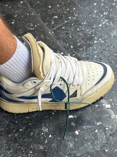 This media file shows a close-up, overhead view of a person's foot wearing a white and blue sneaker with white laces and a green zip tie. The sneaker has a worn appearance, with creasing on the toe box and scuffs on the sole. The sole is a light tan color. The sneaker has blue accents and what appears to be a blue stripe detail. A green zip tie is attached to the laces and wrapped around the shoe, with the excess portion extending outwards. The person is wearing a white ribbed sock, and part of their hairy leg is visible. The background is a dark, textured surface, possibly asphalt or a similar material, with flecks of lighter material embedded in it. The lighting suggests it is daytime, and the scene appears to be outdoors. There is no visible text in the image. The setting is likely an urban or outdoor environment.