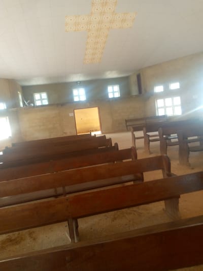 The image depicts the interior of a sparsely furnished church in Madakiya, Nigeria. The view is from a low angle, looking towards the front of the church. Wooden pews, arranged in rows, fill the foreground and middle ground, with their surfaces showing some wear. The walls are a light, earthy color. In the background, several rectangular windows with multiple panes are visible along the walls, letting in bright, natural light that creates glare in some areas of the image. A doorway or entrance is visible in the far wall, leading to a brightly lit space. The ceiling is white and features a large, decorative cross made of square, patterned tiles. The floor appears to be concrete or a similar utilitarian material, with some visible discoloration. No people are visible in the frame. The overall impression is one of a simple, functional place of worship.