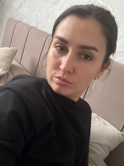 This is a close-up, vertically oriented selfie of a young woman lying down, likely at home in Нижнеяркеево, Russia. She is wearing a black top and has dark hair pulled back. Her face is turned towards the camera, with her eyes looking directly into the lens. She has natural makeup with visible freckles across her nose and cheeks. A small square stud earring is visible in her left ear. The background appears to be a bed or couch with light-colored pillows and a textured, light-colored wall or headboard. The lighting suggests it could be daytime, possibly indoors with natural light. Her expression is relaxed and neutral. No visible text is present in the image.
