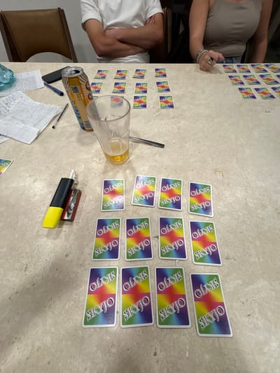 A table shows a game of Skyjo in progress, with cards arranged in grids for multiple players. An empty beer can and half-empty beer glass sit among the cards, alongside a highlighter, a pen, and score sheets. Two people are partially visible at the table, one wearing a white shirt and the other a gray tank top, indicating a casual, relaxed game setting.