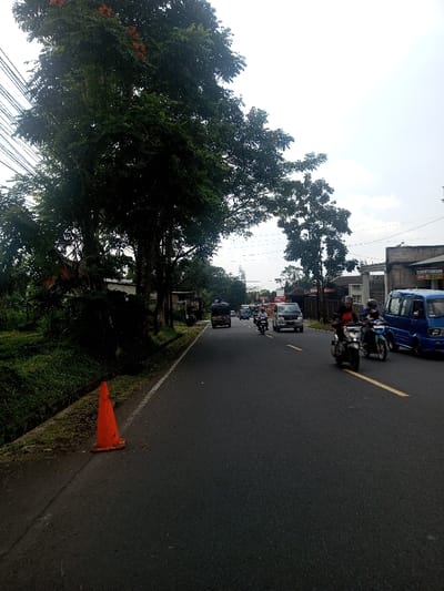 The image captures a daytime scene on a paved road in the city of Niz Lakuk, India, under an overcast sky. In the immediate foreground, an orange traffic cone is positioned on its side on a dirt shoulder to the left of the asphalt roadway, adjacent to a narrow, vegetated drainage ditch. The road extends into the background, marked by a solid yellow line.

Multiple vehicles are present on the road, traveling away from the viewer. In the right lane, a blue compact van is stationary or moving slowly. To its left, two individuals on motorcycles are visible; the rider closer to the center line operates a light-colored scooter, and the rider to their right operates a dark-colored scooter. Both riders wear dark helmets and dark jackets. Further down the right lane, a silver SUV or minivan is present, followed by additional vehicles including a white van and a dark sedan. In the left lane, a dark three-wheeled vehicle, possibly a cargo auto-rickshaw, is heavily loaded with dark material.

The left side of the road is lined with dense green vegetation and several tall trees, some with visible reddish-orange foliage. Utility lines span the upper left quadrant of the frame. On the right side of the road, in the mid-distance, several buildings are visible, including one with a partially obscured sign displaying "TERNAKKAN" in white text on a blue background. The background consists of distant trees, buildings, and a light gray, cloudy sky.