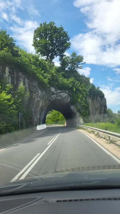 This media file shows a road curving towards a tunnel carved through a rocky, tree-covered mountainside. The road has white double lines and a guardrail on the right. The sky is blue with scattered white clouds, suggesting fair weather. Lush green trees and vegetation cover the slopes surrounding the tunnel. The tunnel entrance is arched and dark, with a hint of sunlight and greenery visible inside. A small white sign with blurry text is visible on the right side of the road near the tunnel entrance. The image appears to be taken from a moving vehicle, as indicated by the perspective and the blurred elements. The overall impression is one of a scenic drive through a natural landscape. The location is Bilka, Bulgaria. There are no people visible in the image. The time of day appears to be daytime.