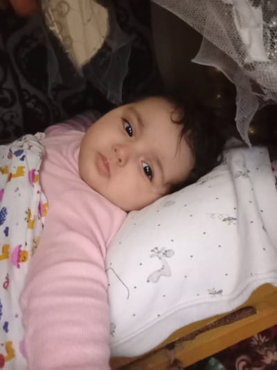 An infant is depicted lying down indoors in what appears to be a bed or crib, located in Galaosiyo, Uzbekistan. The infant has dark hair and dark eyes and is wearing a pink, long-sleeved ribbed shirt. A white blanket with colorful cartoon animal designs (e.g., yellow giraffes, blue patterns) covers the infant's left side. The infant's head rests on a white pillow decorated with grey animal figures (e.g., a giraffe) and small dark dots. The background features dark, patterned fabric, a translucent white mesh material, and some metallic, reflective objects. A wooden edge and a dark, possibly floral-patterned fabric are visible in the lower right portion of the frame. The infant's gaze is directed forward, towards the camera.