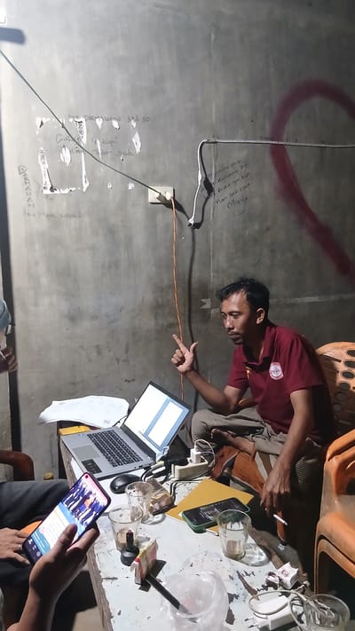 In Keudejungkagajah, Indonesia, a man in a maroon polo shirt with a circular logo is seated on an orange plastic chair, gesturing with his right hand as he looks towards a laptop open on a table. The table is cluttered with two drinking glasses, a pack of cigarettes, a lighter, a smartphone displaying a photo of people, and other assorted items. Another person's hands are visible in the foreground, holding a smartphone. The background is a rough concrete wall with various markings and graffiti. An orange electrical cord runs from a wall socket down to the table. The scene appears to be indoors, likely in a workspace or informal gathering area.