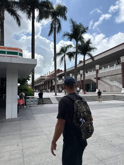 In the city of Xiangzhou, China, a young man wearing a dark t-shirt, black pants, and a navy blue baseball cap walks away from the camera. He is carrying a camouflage backpack with a water bottle visible in a side pocket and what appears to be a "The North Face" and "Supreme" tag on the lower portion. He is walking on a paved plaza in front of a large, multi-story building with an arched entrance and ornate white balustrades. Several palm trees are visible throughout the scene, casting shadows on the plaza. In the background, to the left of the frame, a section of a 7-Eleven convenience store is visible. Other people are walking in the distance, and some are sitting on steps leading up to the building. The sky is partly cloudy with patches of blue.