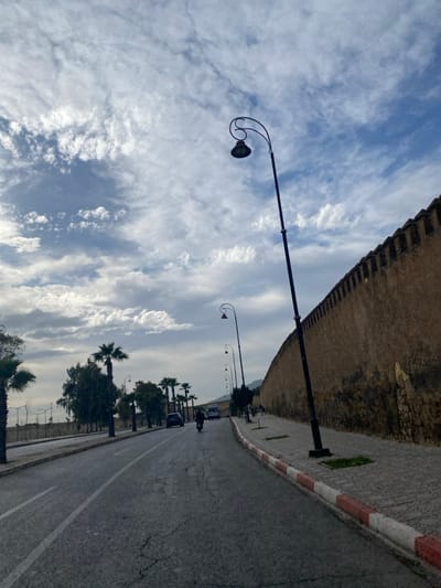 The image presents a daytime street scene in Mechouar Fes Jdid, Morocco, under a partially cloudy sky with patches of blue. A multi-lane asphalt road, exhibiting surface cracks, occupies the central and left portions of the frame. A white solid line marks the left edge of the visible lane, while a faded center line is discernible further down the road.
On the immediate right, a grey paved sidewalk, bordered by a curb painted with alternating red and white stripes, runs parallel to the road. Abutting the sidewalk is a tall, textured ochre-colored wall, characteristic of historical ramparts, extending into the distance with a crenellated top.
Multiple streetlights are visible, featuring dark poles and decorative, curved upper arms supporting the light fixtures. One prominent streetlight in the foreground on the right stands tilted. A line of similar streetlights recedes along the wall. On the left side of the road, a row of palm trees and other deciduous trees are visible behind a low, light-colored barrier or wall.
In the middle ground of the road, a person on a black motorcycle is observed moving away from the viewer. Further down the road on the left, a dark-colored car and a white van are also present. Distant hills are visible on the horizon, partially obscured by atmospheric haze. No other individuals are discernible.