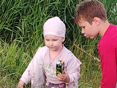 Two children, a young girl and a boy, are standing in a field of tall green grass. The girl, on the left, is wearing a light pink headscarf with a pattern and a light pink long-sleeved shirt with colorful cartoon characters on it. She is holding a small, dark green box in her right hand. She has a necklace around her neck. The boy, on the right, is wearing a bright pink long-sleeved shirt. He is looking down and to his left. The background consists entirely of lush green grass and some dry stalks. The lighting suggests it is daytime, possibly overcast or late afternoon. The scene appears to be outdoors, in a natural environment, potentially a rural area within Заямнаўскі сельскі Савет, Belarus.