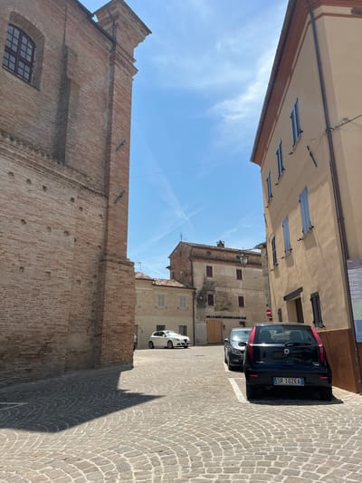 The image shows a narrow street in the town of Barbara, Italy, on a sunny day. The street is paved with cobblestones. On the left is a tall brick building with a large arched window. On the right is a lighter-colored building with blue shutters. Several cars are parked on the street, including a black Fiat Punto in the foreground. In the background, more buildings and a clear blue sky with some wispy clouds are visible. A street sign with Italian text is mounted on the building on the right.