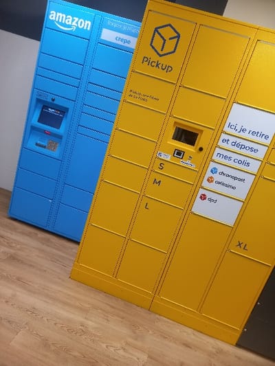 The image displays an indoor scene in Sallaumines, France, featuring two automated parcel locker units situated on a light-colored wooden floor. The unit on the left is a blue Amazon locker, identified by its logo at the top. It incorporates a central interactive screen and a keypad. A white strip above the screen section bears the French text "Bonjour je mange crepe." To its right stands a yellow parcel locker, branded "Pickup" with a cube logo and the clarifying text "Pickup, une offre de La Poste." This yellow unit also features a central screen, an integrated scanner, and compartment sizes labeled S, M, L, and XL. A vertical white panel on its right side reads "Ici, je retire et dépose mes colis" and displays the logos for delivery services Chronopost, Colissimo, and DPD. No individuals are present in the depicted scene.