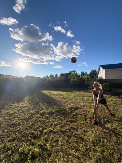A blonde woman wearing a black tank top and dark shorts is playing volleyball in a backyard in Vaskelovo, Russia. The sun is shining brightly, casting long shadows across the dry, mowed grass. A volleyball is in mid-air above her. The background shows houses and trees under a bright blue sky with scattered white clouds.