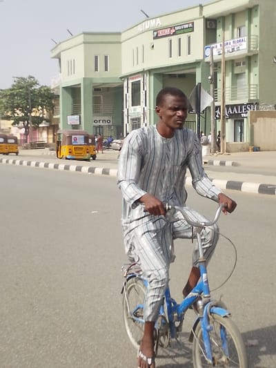 A man is depicted riding a blue bicycle on a paved road in Babban Giji, Nigeria. He is positioned slightly right of center, facing towards the right with his head turned slightly left, looking forward. He wears a light-colored, vertically striped long-sleeved tunic and matching trousers, along with white and black flip-flops. His hands grip the silver handlebars of the blue bicycle, which appears to be a folding type, showing signs of wear and rust.

To the left of the road, a black and white striped curb or median separates the main thoroughfare from a shoulder or parking area. Two yellow motorized tricycles, commonly known as Keke Napep, are visible further down the road on the left, one with a political poster affixed to its rear.

In the background, a large, two-story commercial building with a light green or mint-colored facade dominates the scene. The building features multiple rectangular windows and prominent signage, including "ALTUMA" along the top edge, "KHALIJ TRAVELS & TOURS LTD." on the upper floor, and "BORA GLOBAL" and "AALESH" on the lower level to the right. The word "orno" is visible on the ground floor to the left. Other individuals are present on the sidewalk or near the building in the background. The sky is bright and clear, indicating daylight.