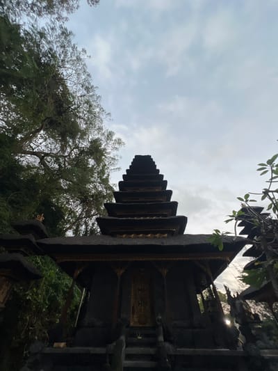 The image shows a tiered, dark stone temple structure with a thatched roof, likely a traditional Balinese Meru temple. The architecture features multiple levels, each with a slightly smaller footprint than the one below, culminating in a pointed top. Ornate golden detailing adorns the edges of the roof layers and a decorative panel on the central doorway. The temple is set amidst lush greenery, with trees and foliage visible around and behind it. The sky above is overcast with clouds. The scene appears to be in the early morning or late afternoon, given the soft lighting. Based on the style of the temple and the surrounding environment, the location is consistent with Dawan, Indonesia, known for its traditional temples and cultural heritage.