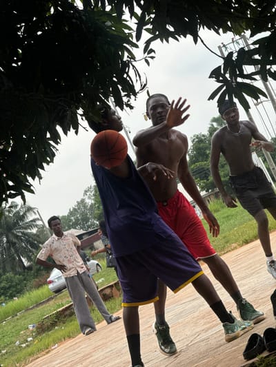 In Amansee, Nigeria, several young men are engaged in a game of basketball on an outdoor court. The court is made of a reddish-brown material, with dirt and grass bordering it. Overhead, the branches of a large tree with dark green leaves cast shadows across the scene.
The central figures are two shirtless young men, one in a purple tank top and purple shorts with a yellow trim, holding an orange basketball. He is in the process of shooting or passing the ball. The other player, wearing red shorts, is positioned to defend. Another shirtless young man in black shorts stands further back, observing the game.
In the background, a man in a patterned short-sleeved shirt and gray trousers stands with his hands on his hips, seemingly a spectator. A silver sedan is parked on the grassy area behind him. The sky is overcast, contributing to the diffused lighting. Scattered pieces of litter are visible on the ground around the court. The overall impression is a casual, energetic game of basketball taking place in a rural or semi-urban setting within Amansee.