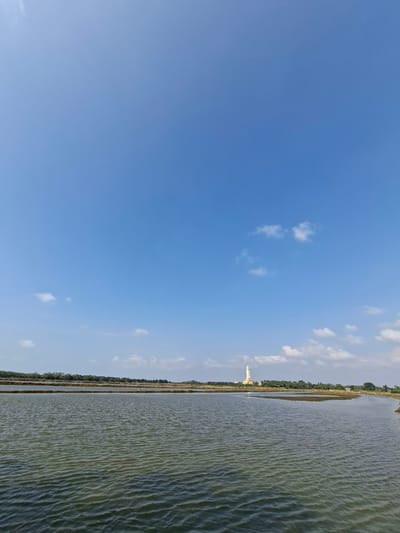 The image displays a vast expanse of water on the right side and a clear blue sky on the left. A thin strip of land with trees and a tall white structure, possibly a minaret or a tower, runs along the horizon separating the water from the sky. The water surface is rippled, suggesting a light breeze. A few scattered white clouds dot the sky. The lighting indicates it is daytime, likely late morning or afternoon, with bright sunlight. The scene appears to be a rural or natural landscape, possibly near a body of water such as a lake, river, or coastal area. The presence of the tall structure hints at a settlement or a place of worship. There is no visible text in the image.
