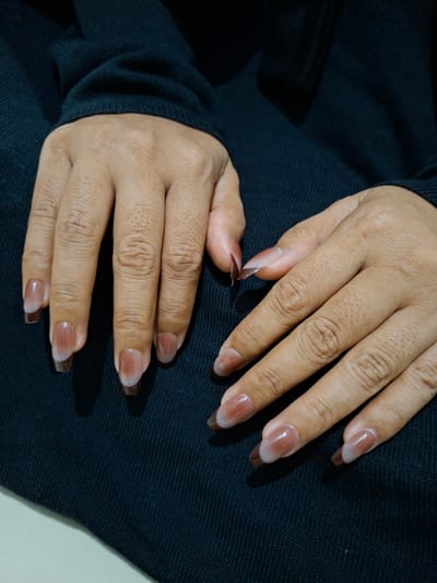 The image shows a close-up of two hands with manicured nails. The nails are clear at the base and transition to a brown color at the tips, in a rounded shape. The hands are resting on a dark navy blue knit fabric, possibly a sweater or blanket. The skin tone of the hands is tan. The lighting is somewhat dim, with shadows visible on the hands and fabric. The background appears to be a plain light surface.
